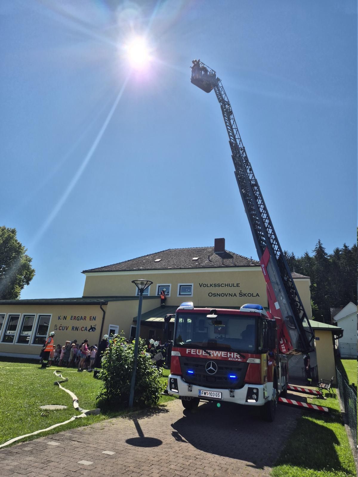 Ein Feuerwehrauto mit ausgefahrener Leiter steht an einem sonnigen Tag vor einem Schulgebäude.