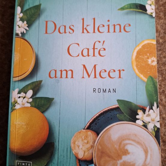 Buchcover von 'Das kleine Cafe am Meer' mit einer Orange, Kaffee und Blumen auf blauem Hintergrund.