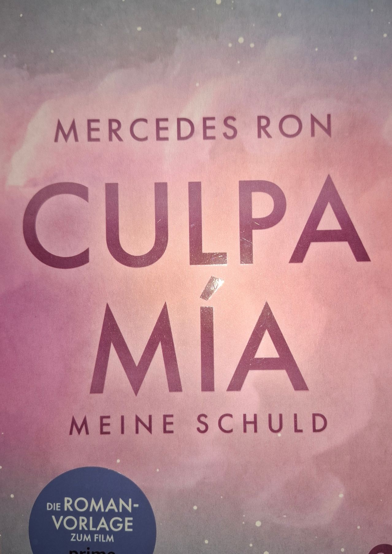 Buchcover von 'Culpá Mía' von Mercedes Ron. Es hat einen rosa Hintergrund und lila Text. 'Culpá Mía' bedeutet auf Englisch 'Meine Schuld'.