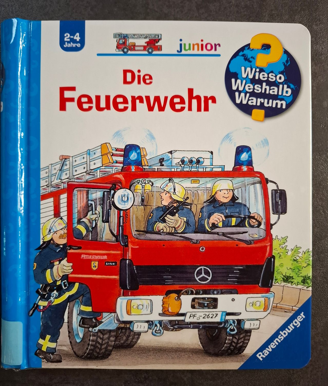 Buchcover für Kinder im Alter von 2-4 Jahren mit dem Titel 'Die Feuerwehr', das einen roten Feuerwehrwagen mit Feuerwehrleuten im Inneren und einem Feuerwehrmann außerhalb zeigt. Das Cover hat einen Globus mit einem Fragezeichen und dem Text 'Wieso Weshalb Warum'.