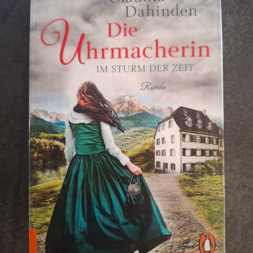 Das Buchcover zeigt eine Frau in einem grünen Kleid, die auf einem Kopfsteinpflasterweg geht, mit einem Haus im Hintergrund. Der Titel ist 'Die Uhrmacherin' und der Autor ist Claudia Dahinden.