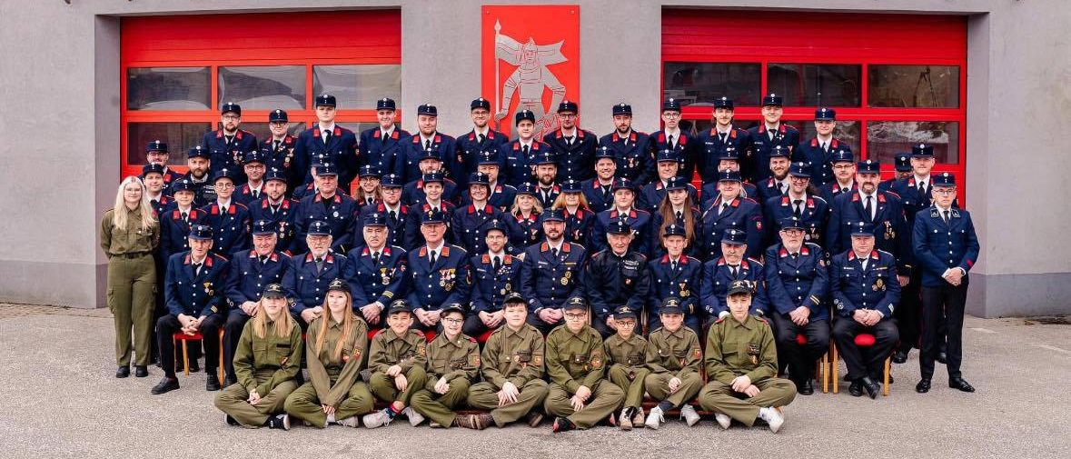 Eine große Gruppe uniformierter Personen, möglicherweise Feuerwehrleute, posiert für ein Foto vor einem Feuerwehrhaus mit roten Türen.