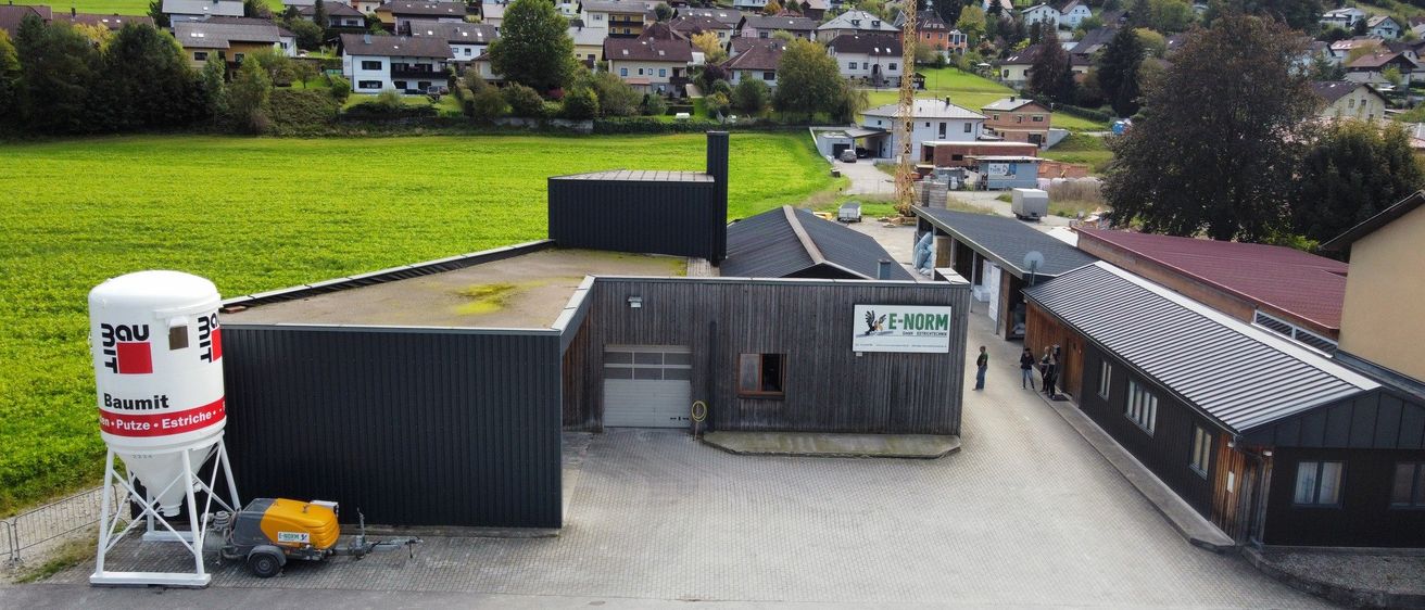 Luftaufnahme eines Bauernhofs mit einem großen Gebäude mit der Aufschrift E-NORM, einem Kran und Wohnhäusern im Hintergrund, umgeben von grünen Feldern und Bäumen.