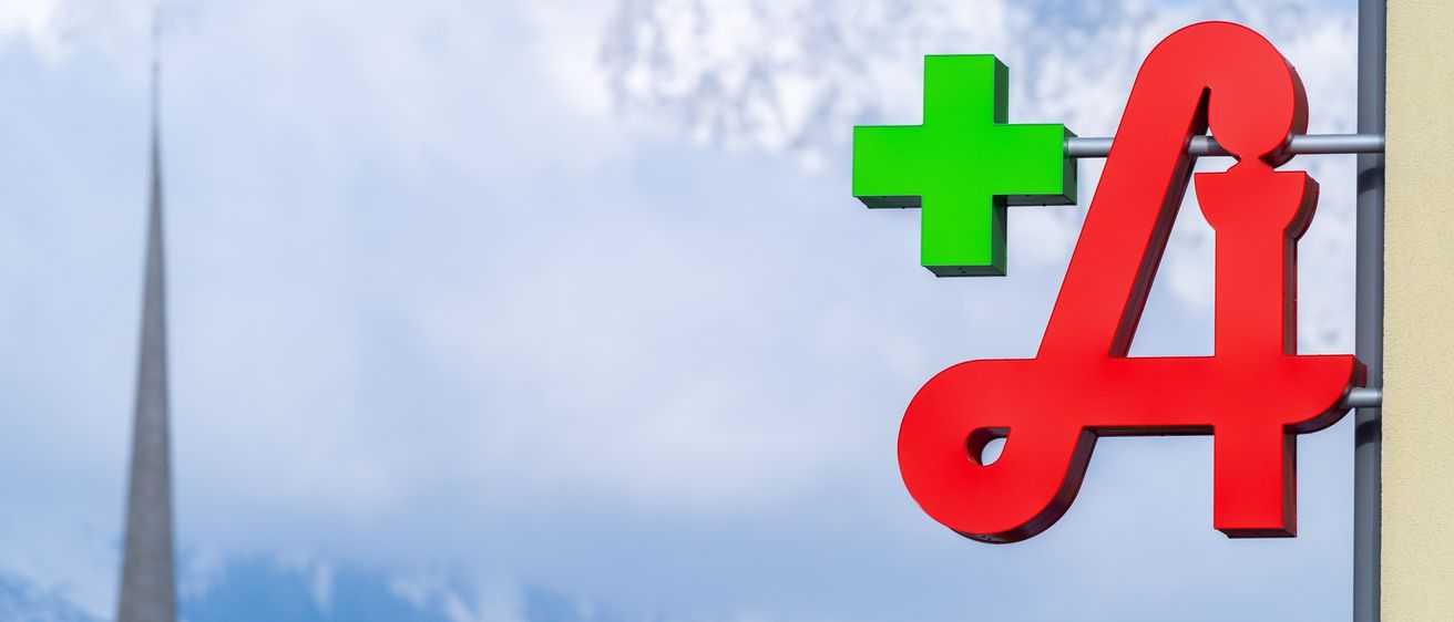 Ein rotes A-Schild mit einem grünen Pluszeichen auf der linken Seite steht vor einem Hintergrund aus verschneiten Bergen unter einem blauen Himmel.