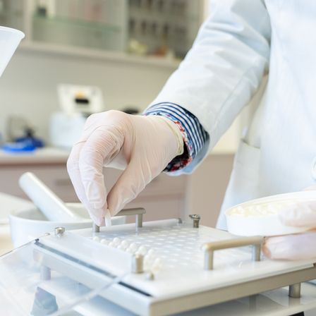 Eine Person in Laborkittel und Handschuhen greift nach kleinen weißen Perlen auf einem Labortablett.