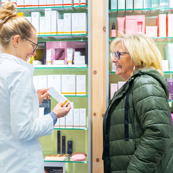 Eine Frau in einem weißen Mantel spricht mit einer anderen Frau in einer grünen Jacke in einer Apotheke.