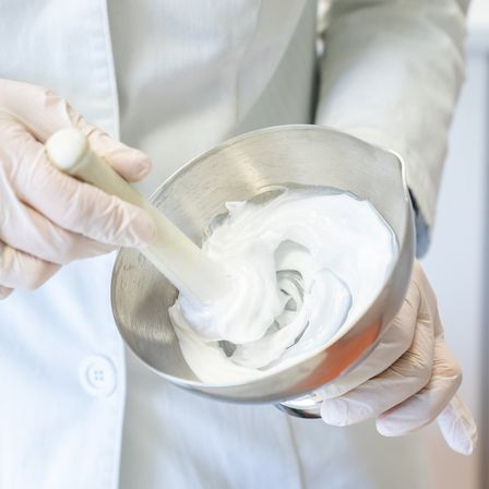 Eine Person in Laborkittel und Handschuhen rührt mit einem Holzlöffel eine weiße Substanz in einer Metallschüssel um.