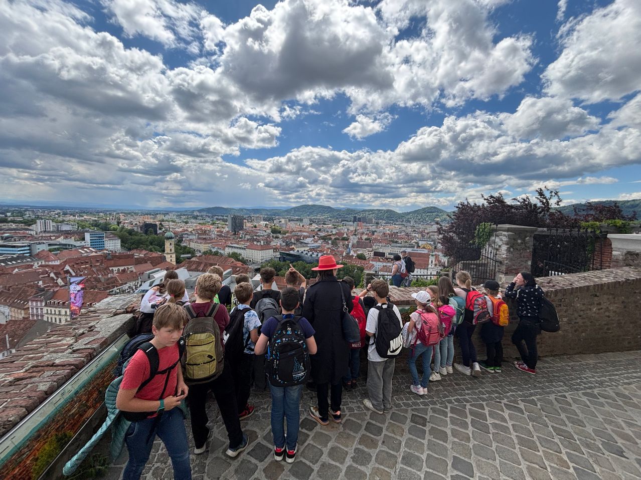 Eine Gruppe von Schulkindern betrachtet eine Stadtlandschaft unter einem bewölkten Himmel von einem hohen Aussichtspunkt aus. Die Gebäude sind rot, und Berge sind in der Ferne sichtbar.