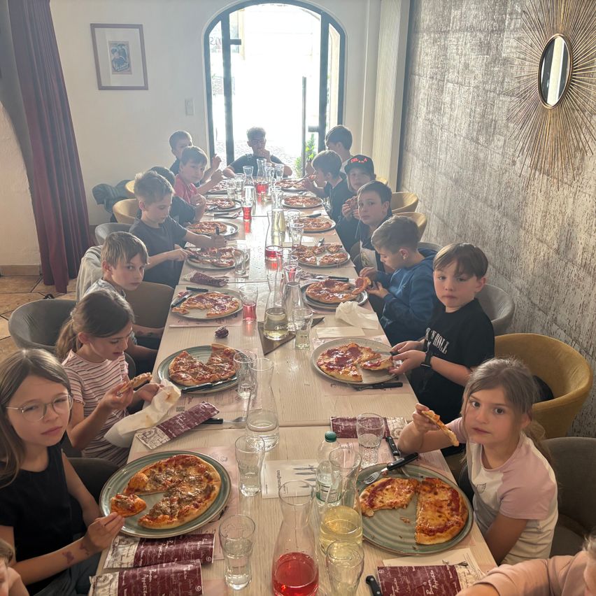 Eine Gruppe von Kindern sitzt um einen langen Tisch herum und isst Pizza. Der Tisch ist mit Tellern, Gläsern und Servietten gefüllt. Die Kinder schauen in die Kamera.