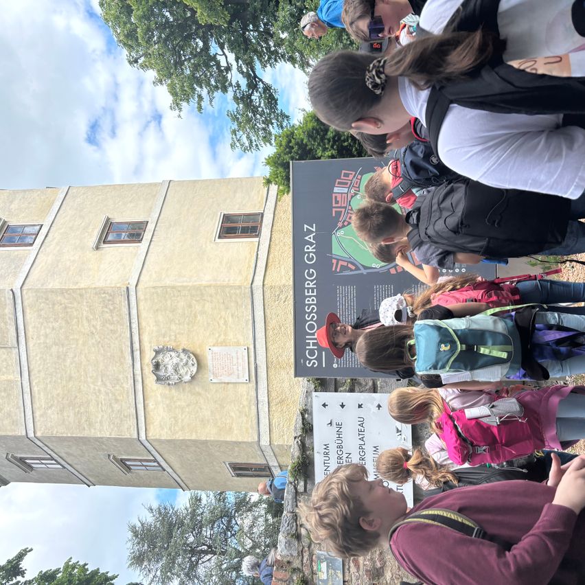 Touristen stehen vor Schlossberg Graz, einige tragen Hüte und haben Rucksäcke. Das Gebäude hat mehrere Fenster und ein Schild.