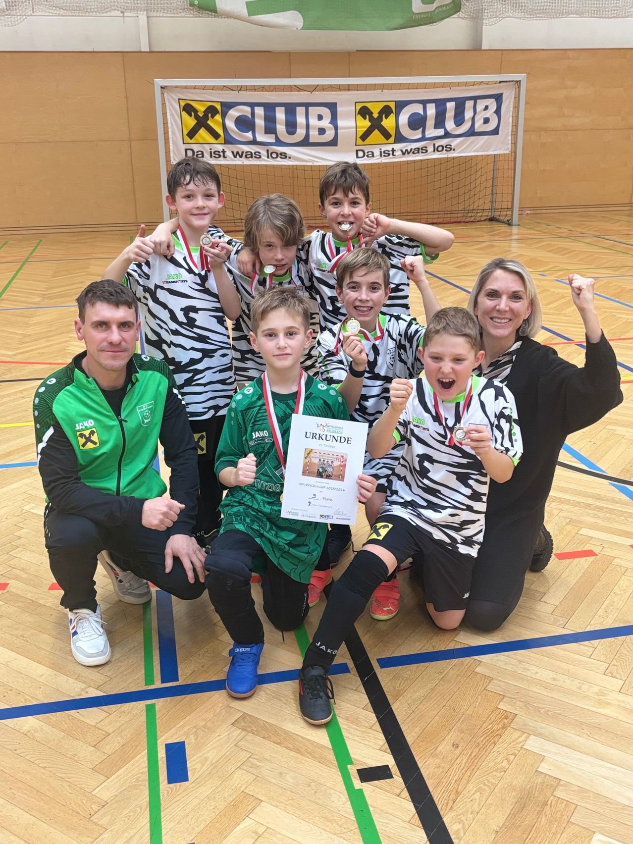 Eine Gruppe junger Athleten mit Medaillen und einem Zertifikat posiert für ein Foto mit einer Frau und einem Mann in einer Turnhalle.