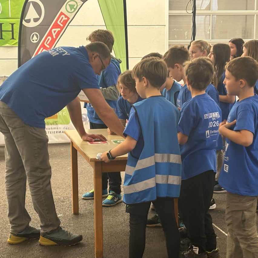A group of children with blue shirts gather around a table where a man explains something to them. Behind the man is a banner.