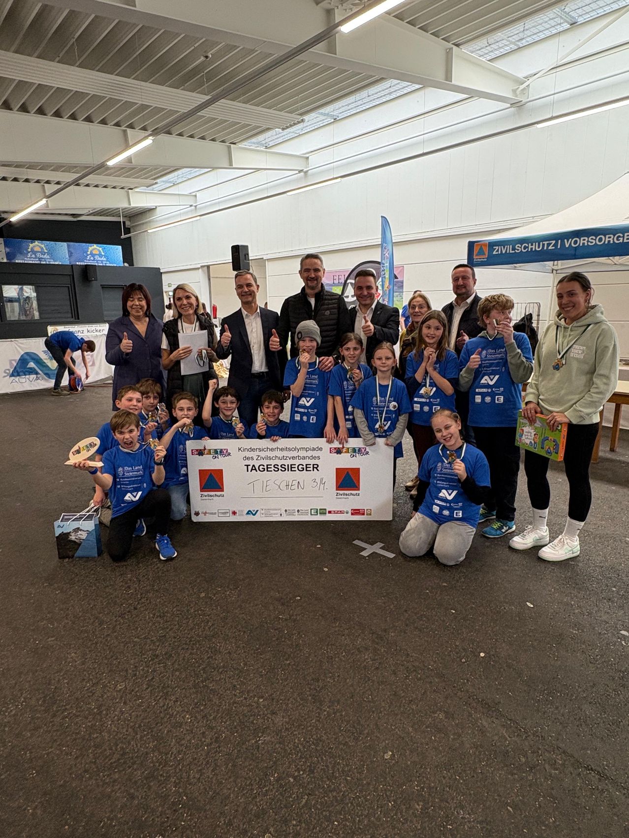 A group of children and adults are gathered, wearing blue shirts with logos, posing for a photo. They hold a certificate and medals. A person bends down. Behind them is a table with a book and a banner.