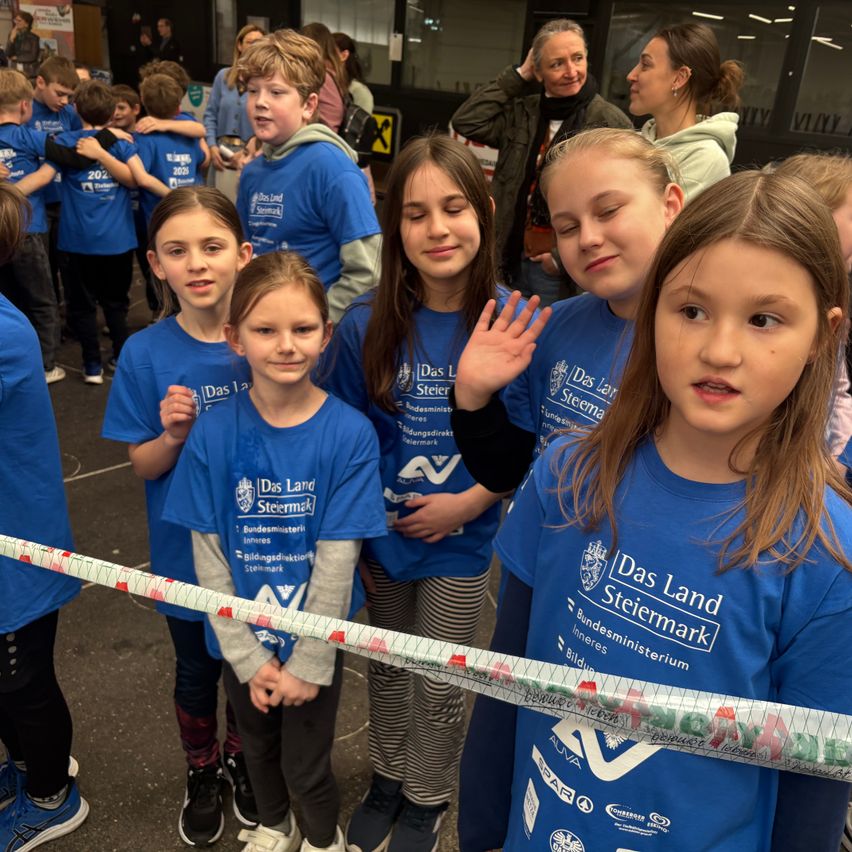 A group of children and adults wearing blue shirts with 'Das Land Steiermark' are standing in front of a building. Some children are holding a long, colorful ribbon.