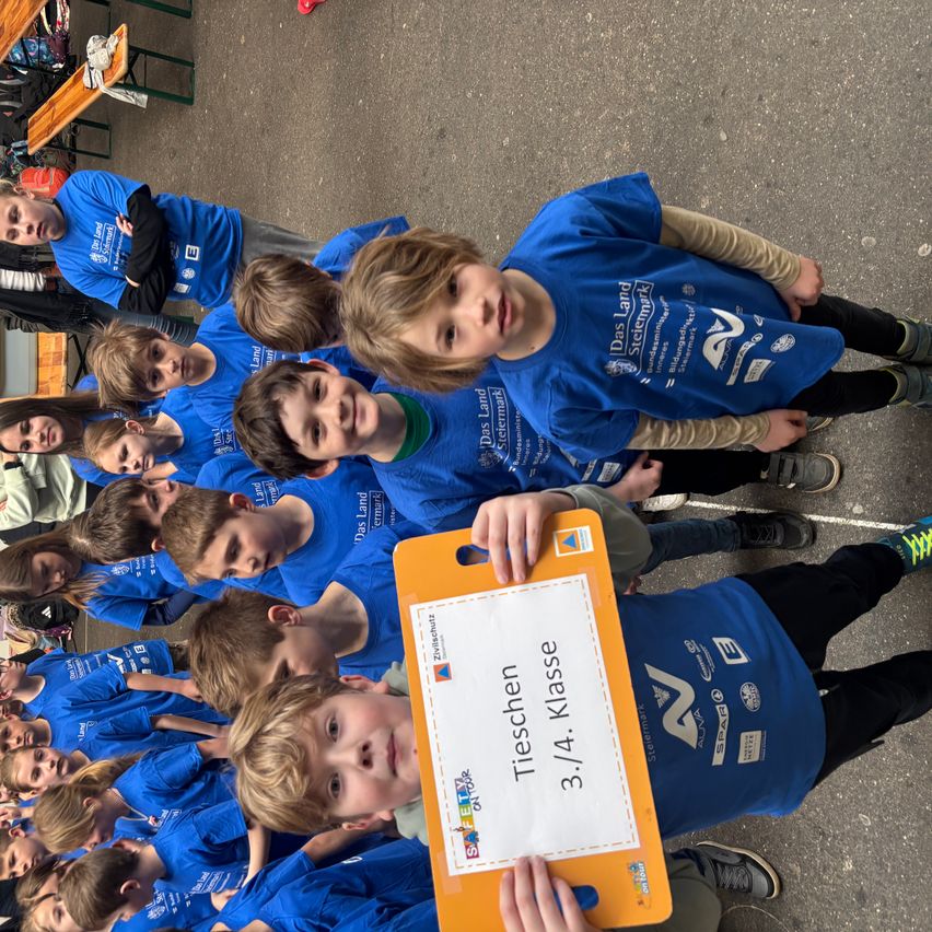 A group of children wearing blue shirts with logos are standing and sitting on the ground. One child is holding a sign with the text 'Tischen 3./4. Klasse'. They are probably at a school event.