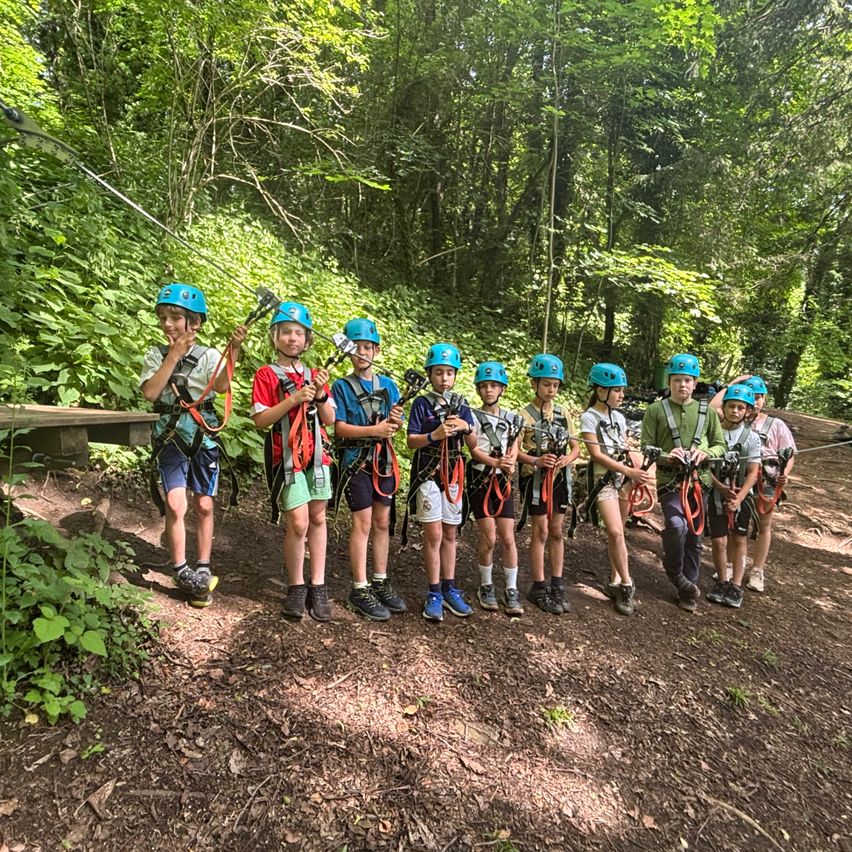 Eine Gruppe von Kindern mit Helmen und Gurten steht in einem Wald und bereitet sich wahrscheinlich auf eine Abenteueraktivität vor.