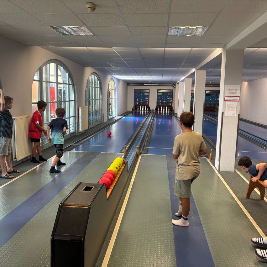 Mehrere Kinder spielen Bowling in einer Indoor-Anlage. Ein Kind wartet an der Bahn auf seinen Zug.