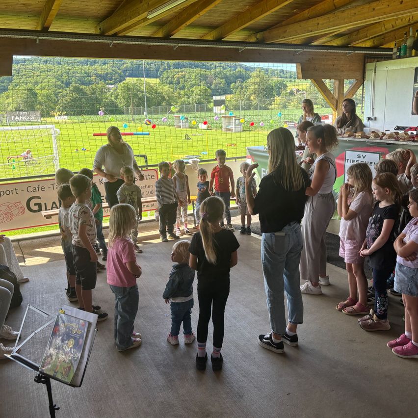 Eine Gruppe von Kindern und Erwachsenen versammelt sich in einem offenen Bereich mit Blick auf ein Fußballfeld. Einige stehen, andere sitzen. Eine Frau steht vor einem Tisch mit Essen und Getränken.