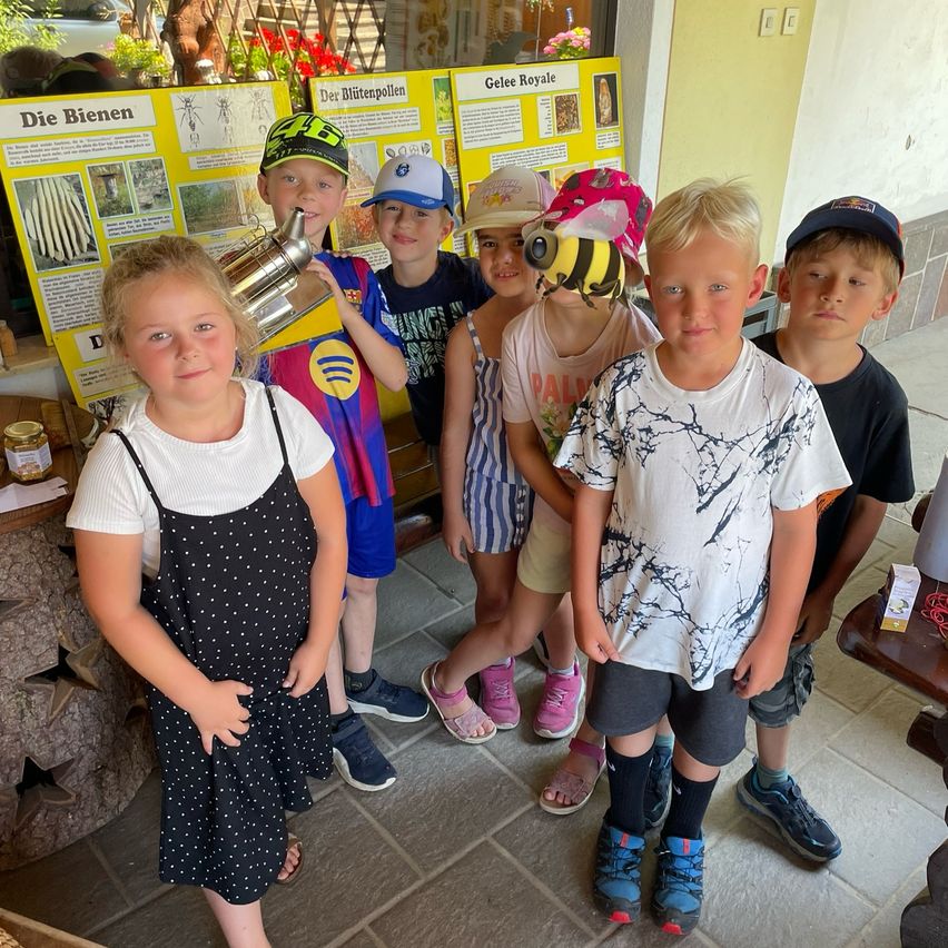 Mehrere Kinder posieren für ein Foto, während sie Mützen tragen, einige mit Bienendesigns, vor einem Lehrposter.