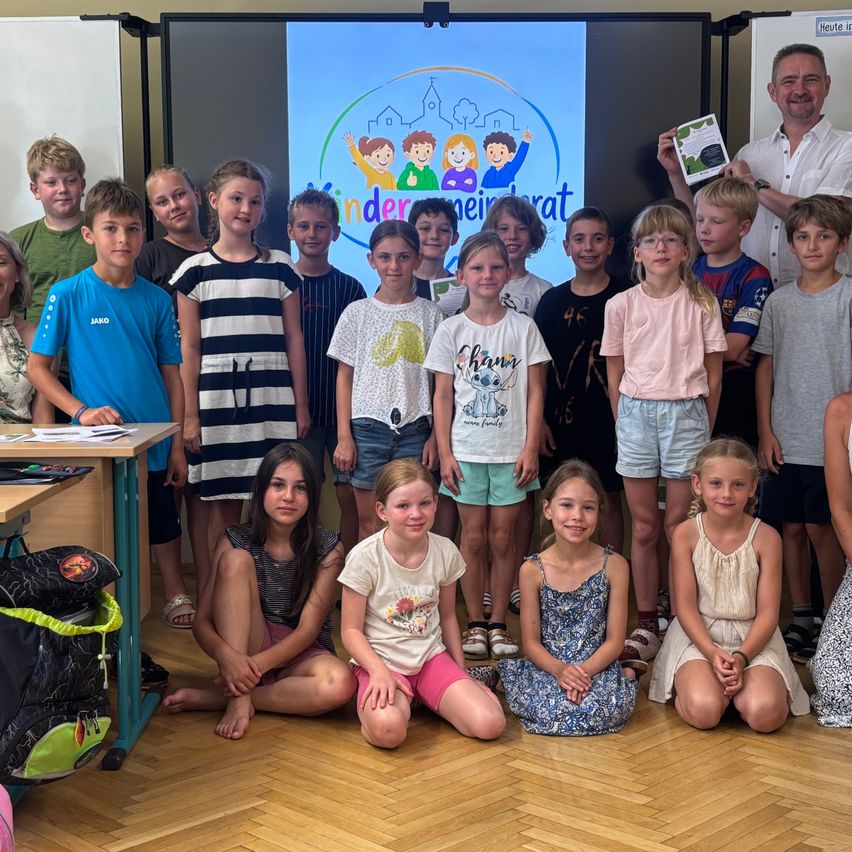 Ein Klassenzimmer mit Kindern und einem Lehrer. Ein Mann hält ein Zertifikat, und Kinder sitzen und stehen vor einem Bildschirm, der ein Bild zeigt.