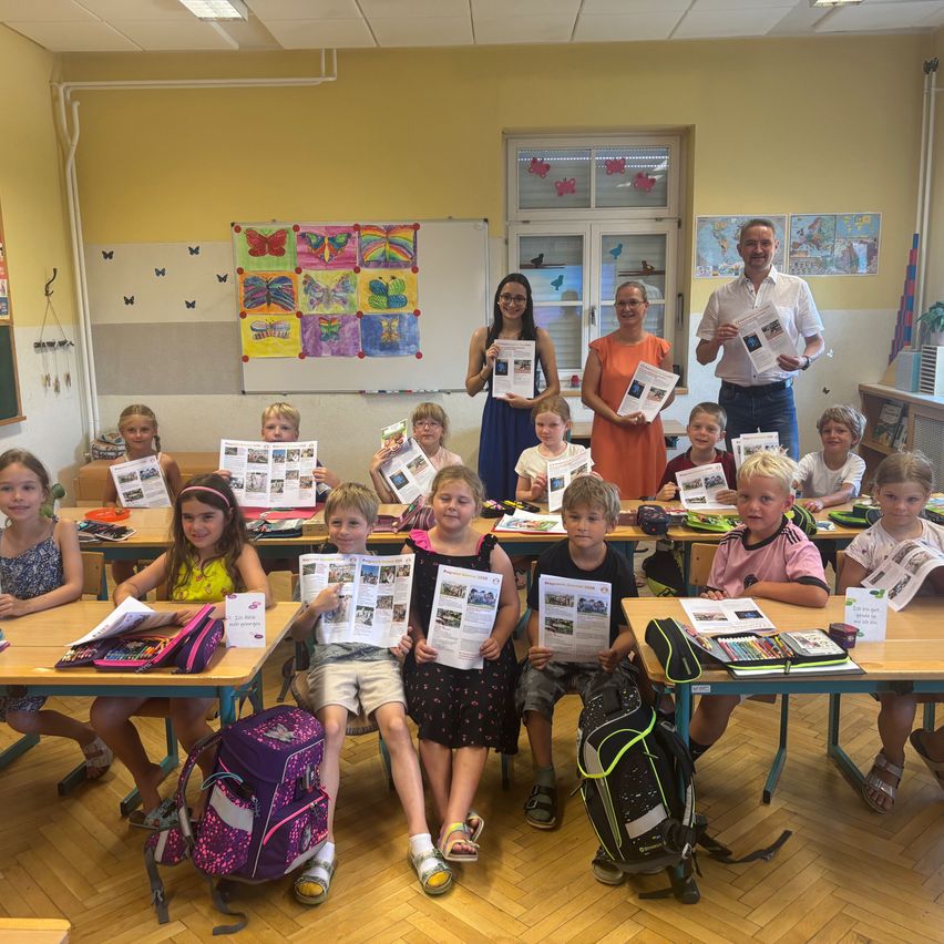 Ein Klassenzimmer voller Kinder und Lehrer, die Bücher halten und für ein Foto lächeln. Der Raum hat Holzböden und gelbe Wände. Zwei Lehrer stehen im Hintergrund.