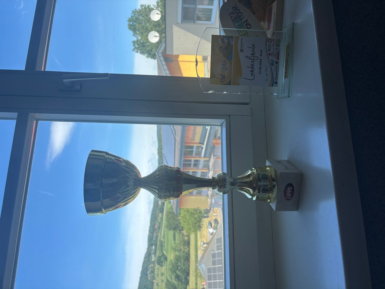 Ein Pokal steht auf der Fensterbank mit freier Sicht aufs Freie. Der Pokal hat einen goldenen Finish und das Wort Lippe darauf. Hinter dem Pokal ist ein durchsichtiger Stand mit einem Zertifikat.