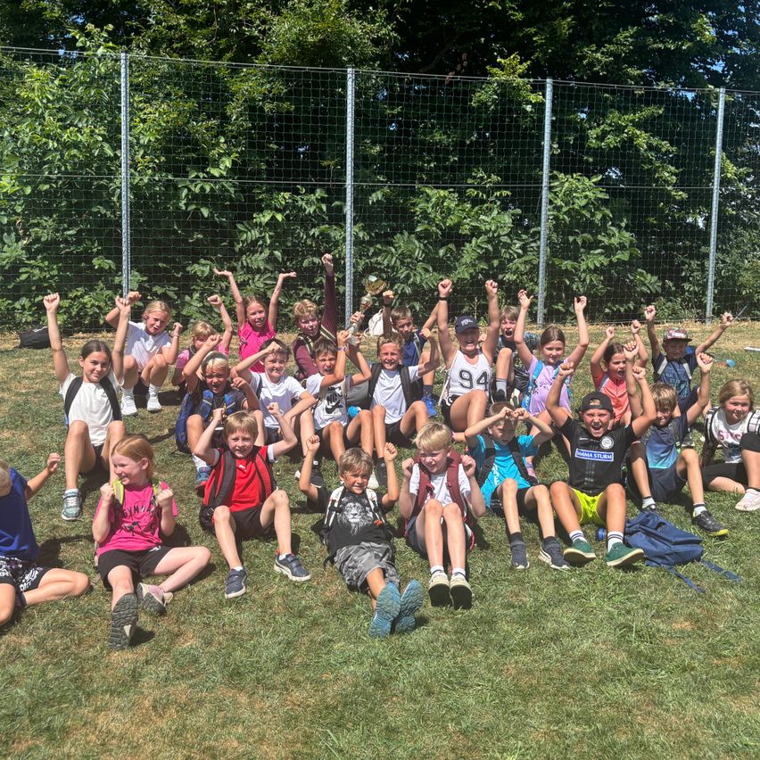 Eine Gruppe von Kindern sitzt und hockt auf dem Rasen und posiert für ein Foto mit erhobenen Händen. Sie tragen verschiedene Kleidungsstücke und Turnschuhe. Dahinter befindet sich ein Metallzaun mit Bäumen.