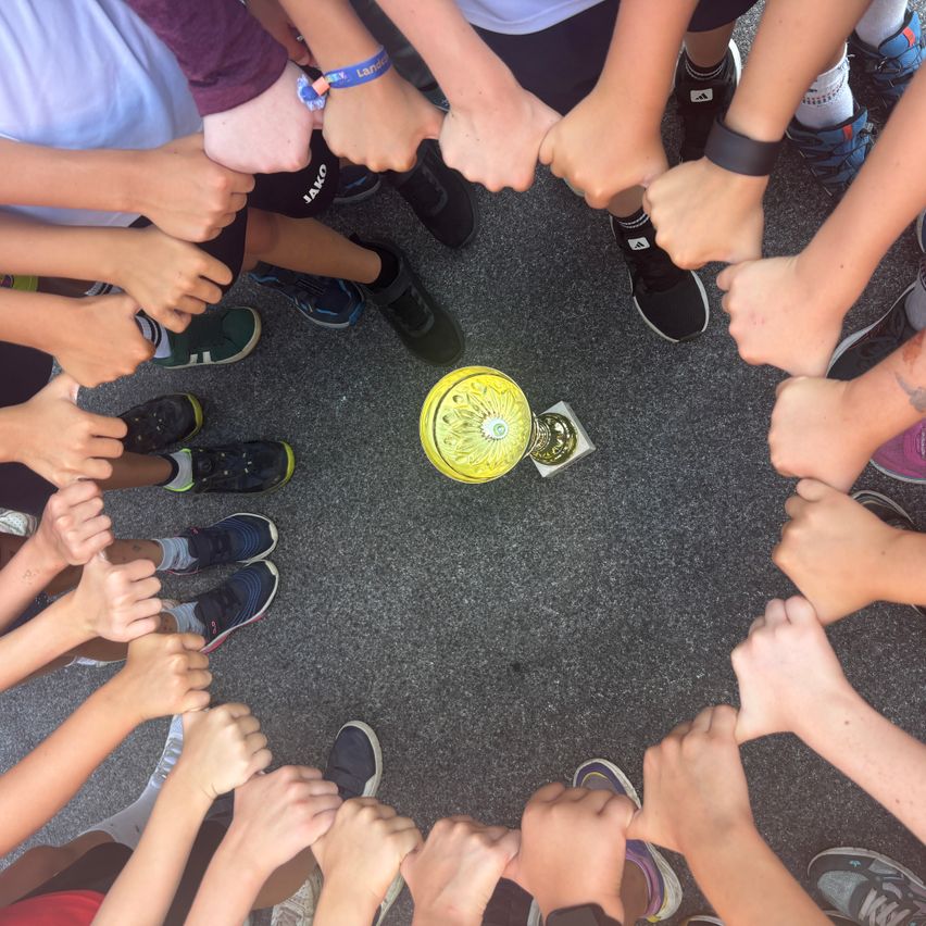 Gruppe von Kindern steht im Kreis um einen gelben Pokal mit verschränkten Händen, trägt Turnschuhe und Armbänder.