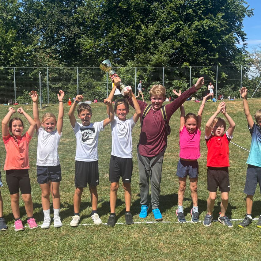 Eine Gruppe von Kindern und zwei Erwachsenen steht auf einem Feld, hebt die Hände und lächelt. Sie tragen Sportkleidung und Turnschuhe. Ein Kind hält einen Pokal. Hinter ihnen trennt ein Zaun das Feld von einem Grasbereich mit Bäumen.