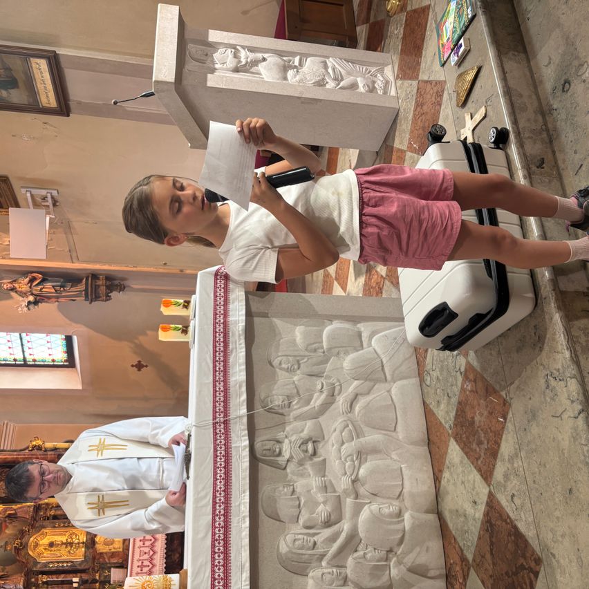 Ein Mädchen steht mit einem Mikrofon an einer Kanzel und liest ein Papier in einer Kirche. Ein Priester kniet vor ihr. Die Kanzel hat eine geschnitzte Steinskulptur dahinter.