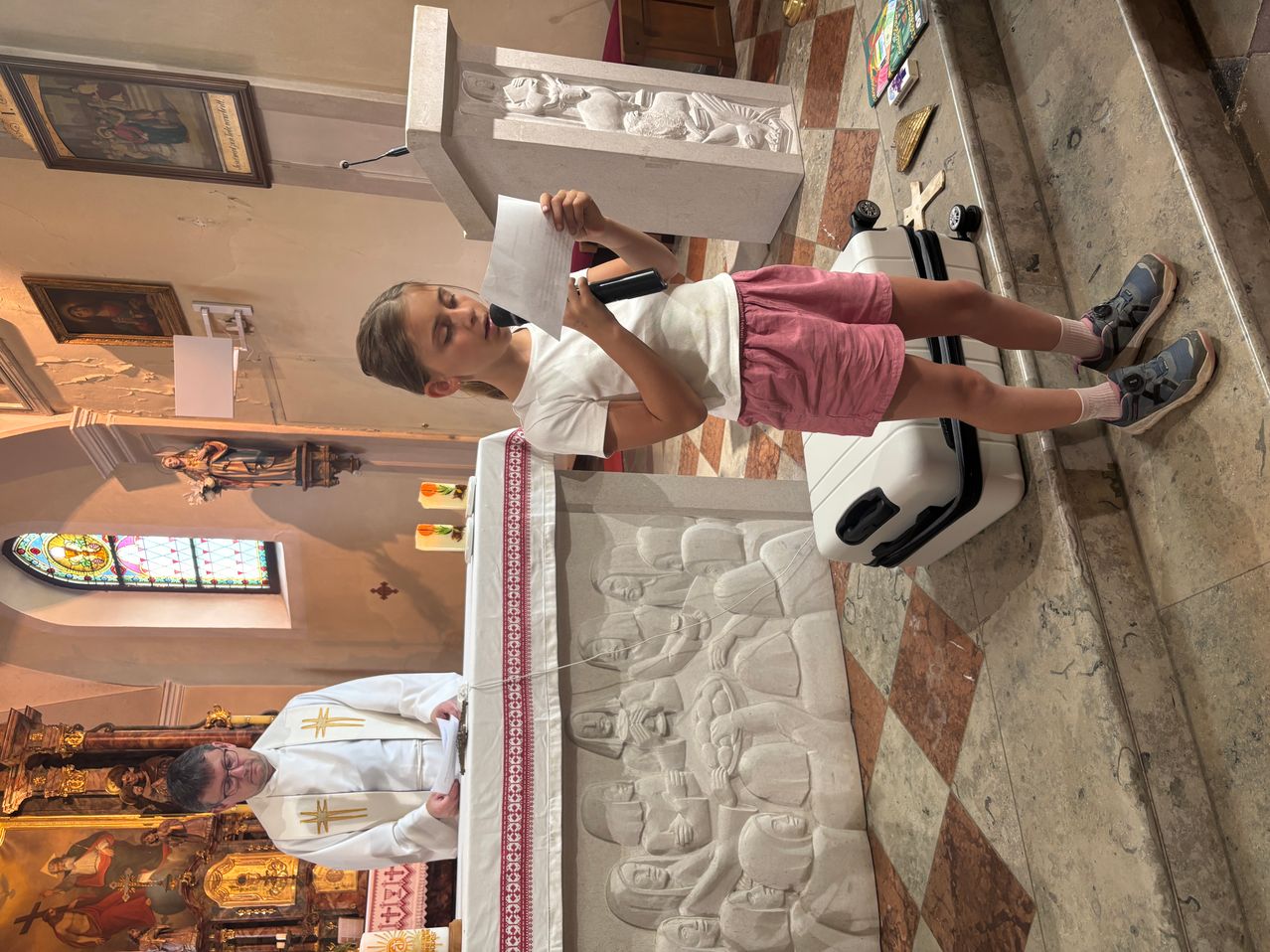 Ein Mädchen steht mit einem Mikrofon an einer Kanzel und liest ein Papier in einer Kirche. Ein Priester kniet vor ihr. Die Kanzel hat eine geschnitzte Steinskulptur dahinter.