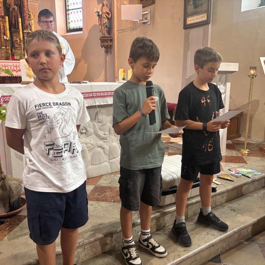 Drei junge Jungen stehen auf den Stufen einer Kirche. Einer hält ein Mikrofon und die anderen zwei halten Papiere. Dahinter steht ein Altar mit einem Priester.
