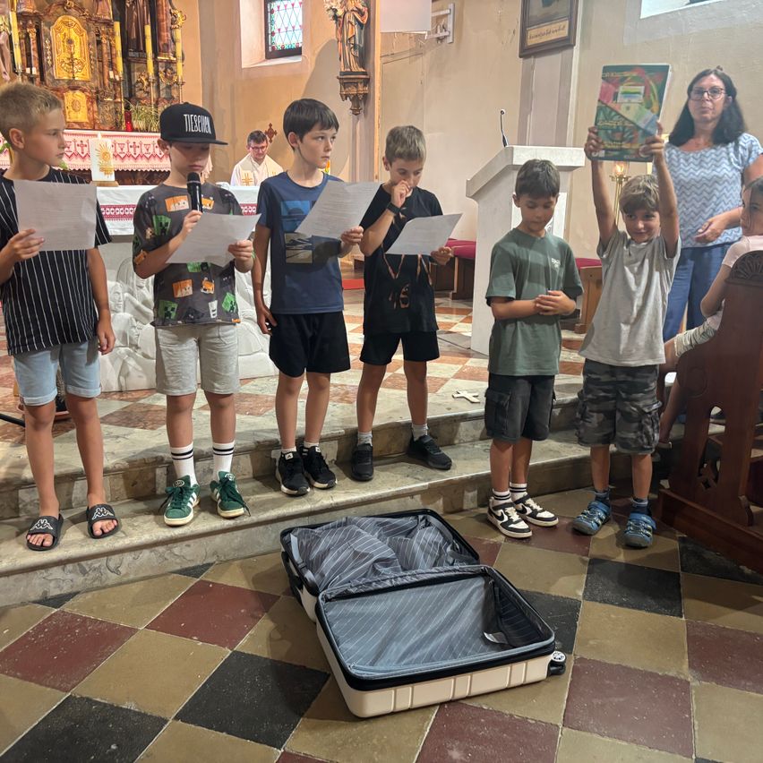 Sechs Jungen halten Papiere und einer ein Buch, stehen in einer Kirche mit einem Priester in der Nähe. Ein offener Koffer liegt auf dem Boden.