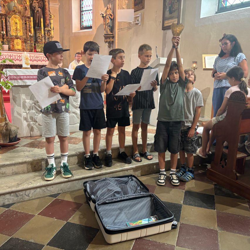 Eine Gruppe von Kindern steht in einer Kirche und hält Papiere. Ein Koffer liegt offen auf dem Boden.