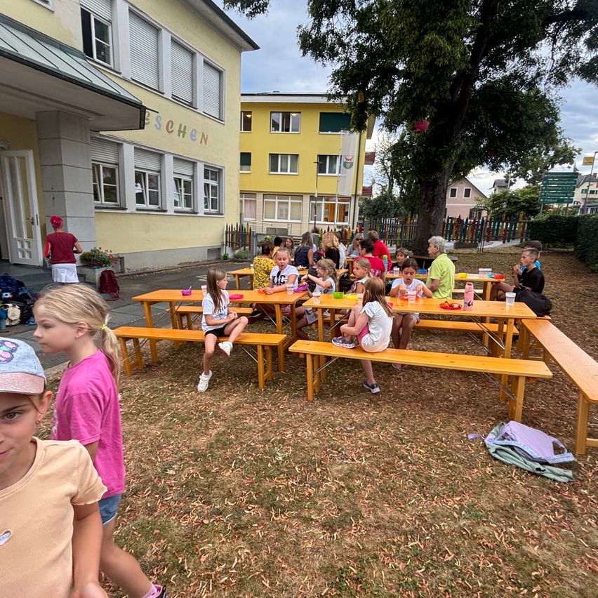 Mehrere Kinder essen an einem Picknicktisch vor einem gelben Gebäude. Eine Frau steht in der Nähe des Eingangs. Im Hintergrund befinden sich Bäume, Pflanzen und ein Zaun.