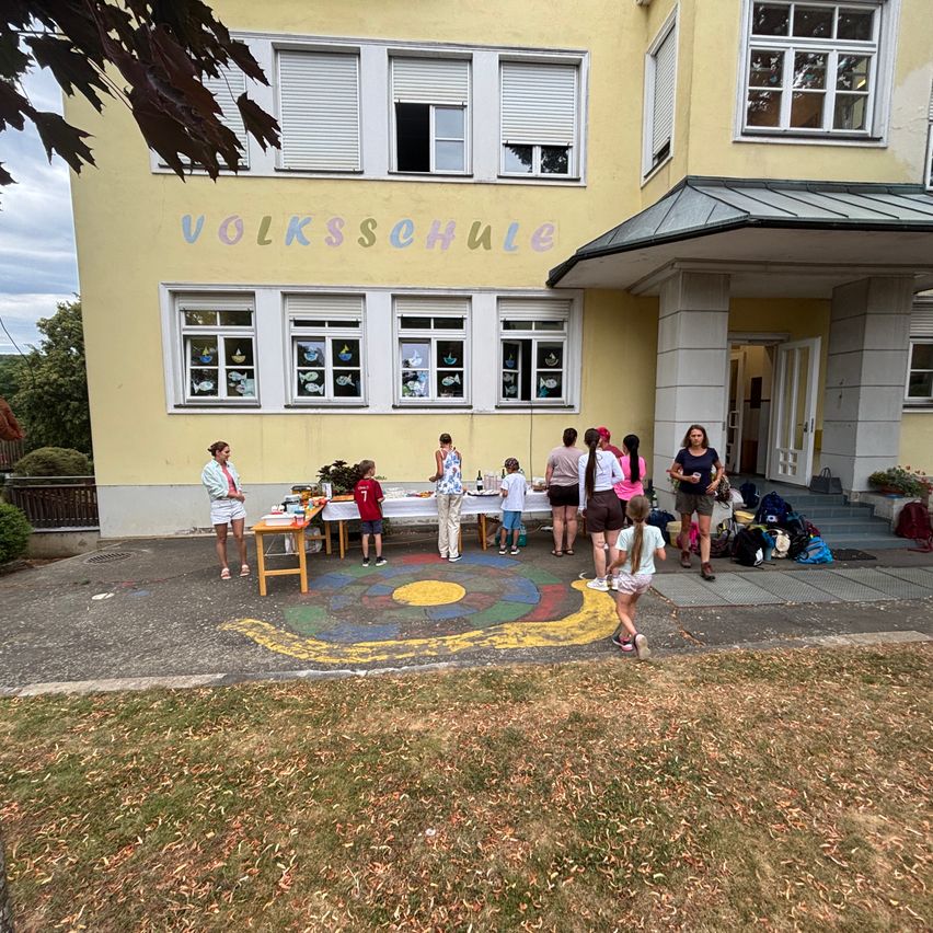 Mehrere Menschen versammeln sich vor einem gelben Gebäude mit dem Wort Volksschule. Einige stehen und andere sitzen. Sie scheinen ein Picknick zu machen. Ein farbenfroher Kreis liegt auf dem Boden.