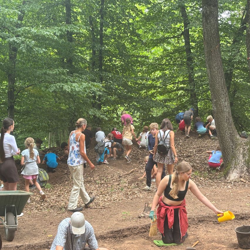 Eine Gruppe von Menschen, einschließlich Kindern, gräbt in einem Wald. Einige tragen Hüte, Brillen und Turnschuhe. Eine Frau hält eine gelbe Kelle und eine Schaufel. Bäume und ein Schubkarren sind in der Nähe.