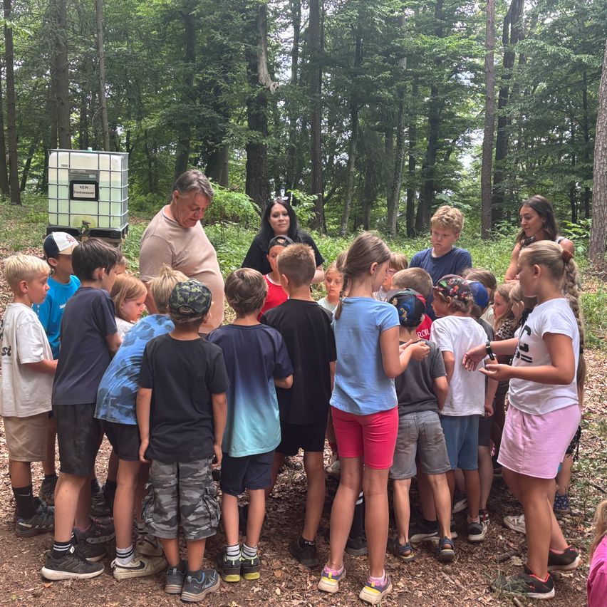 Eine Gruppe von Kindern und Erwachsenen ist in einem Wald versammelt. Sie schauen einem Mann zu, der zu ihnen spricht. Die Kinder tragen Hüte, Turnschuhe und einige haben Armbänder. Hinter ihnen steht ein Tank mit grünem Deckel.