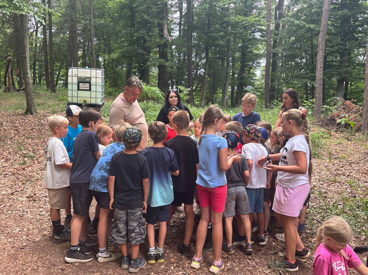 Eine Gruppe von Kindern und Erwachsenen ist in einem Wald versammelt. Sie schauen einem Mann zu, der zu ihnen spricht. Die Kinder tragen Hüte, Turnschuhe und einige haben Armbänder. Hinter ihnen steht ein Tank mit grünem Deckel.