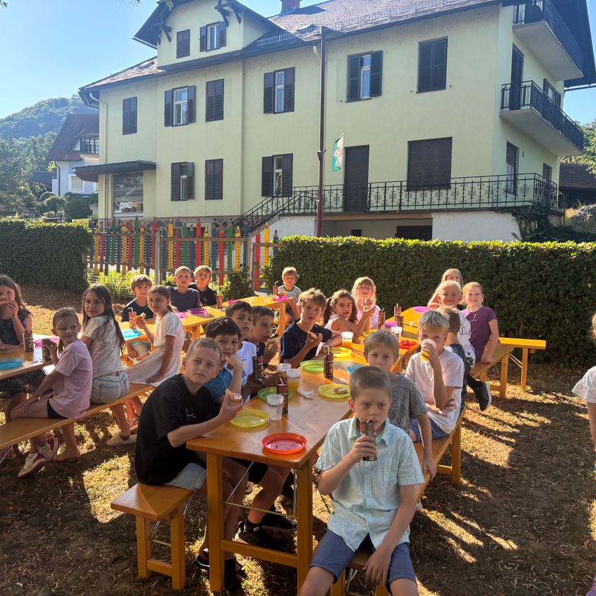 Eine Gruppe von Kindern sitzt an einem langen Tisch im Hof, isst und trinkt. Dahinter steht ein Gebäude mit vielen Fenstern und einer Fahne.