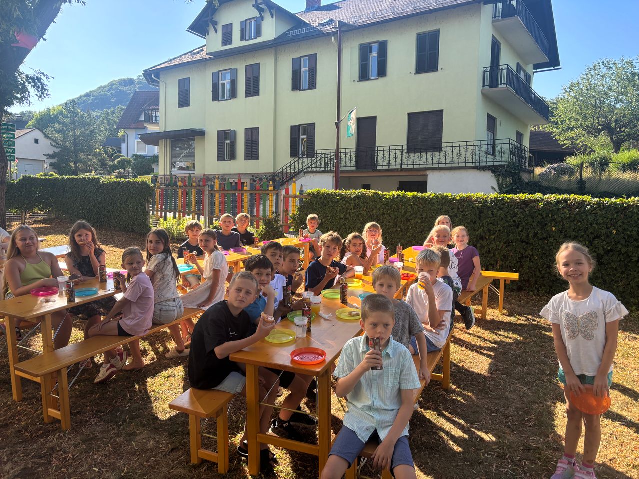Eine Gruppe von Kindern sitzt an einem langen Tisch im Hof, isst und trinkt. Dahinter steht ein Gebäude mit vielen Fenstern und einer Fahne.