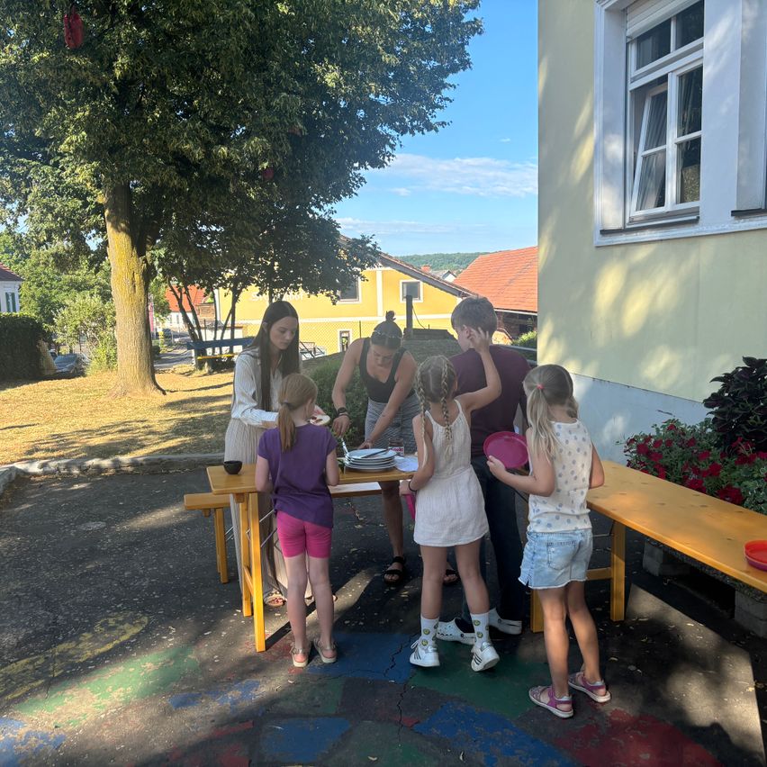 Eine Gruppe von Kindern und Erwachsenen versammelt sich um einen Tisch im Garten und bereitet Essen zu. Der Tisch ist mit Tellern und Schüsseln gedeckt. Die Kinder tragen Freizeitkleidung.