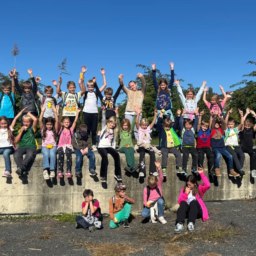 Eine Gruppe von Kindern, einige auf einem Betonwall sitzend, andere stehend, alle mit erhobenen Händen, lächeln für ein Foto unter einem klaren blauen Himmel.
