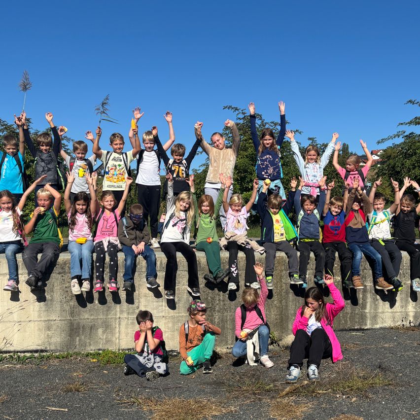 Eine Gruppe von Kindern, einige auf einem Betonwall sitzend, andere stehend, alle mit erhobenen Händen, lächeln für ein Foto unter einem klaren blauen Himmel.