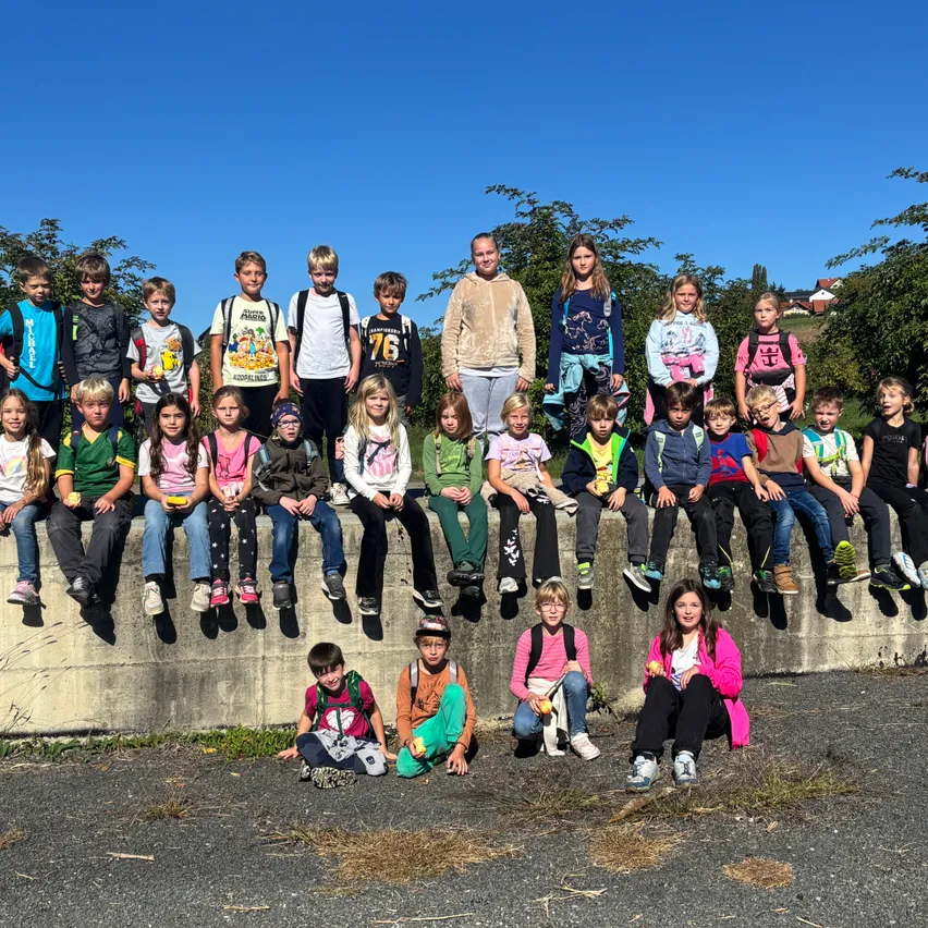 Eine Gruppe von Kindern, einige mit Rucksäcken, sitzen auf einer Betonbarriere und posieren für ein Foto. Die Kinder tragen verschiedene farbige Hemden und Turnschuhe. Der Hintergrund zeigt Bäume und einen klaren blauen Himmel.