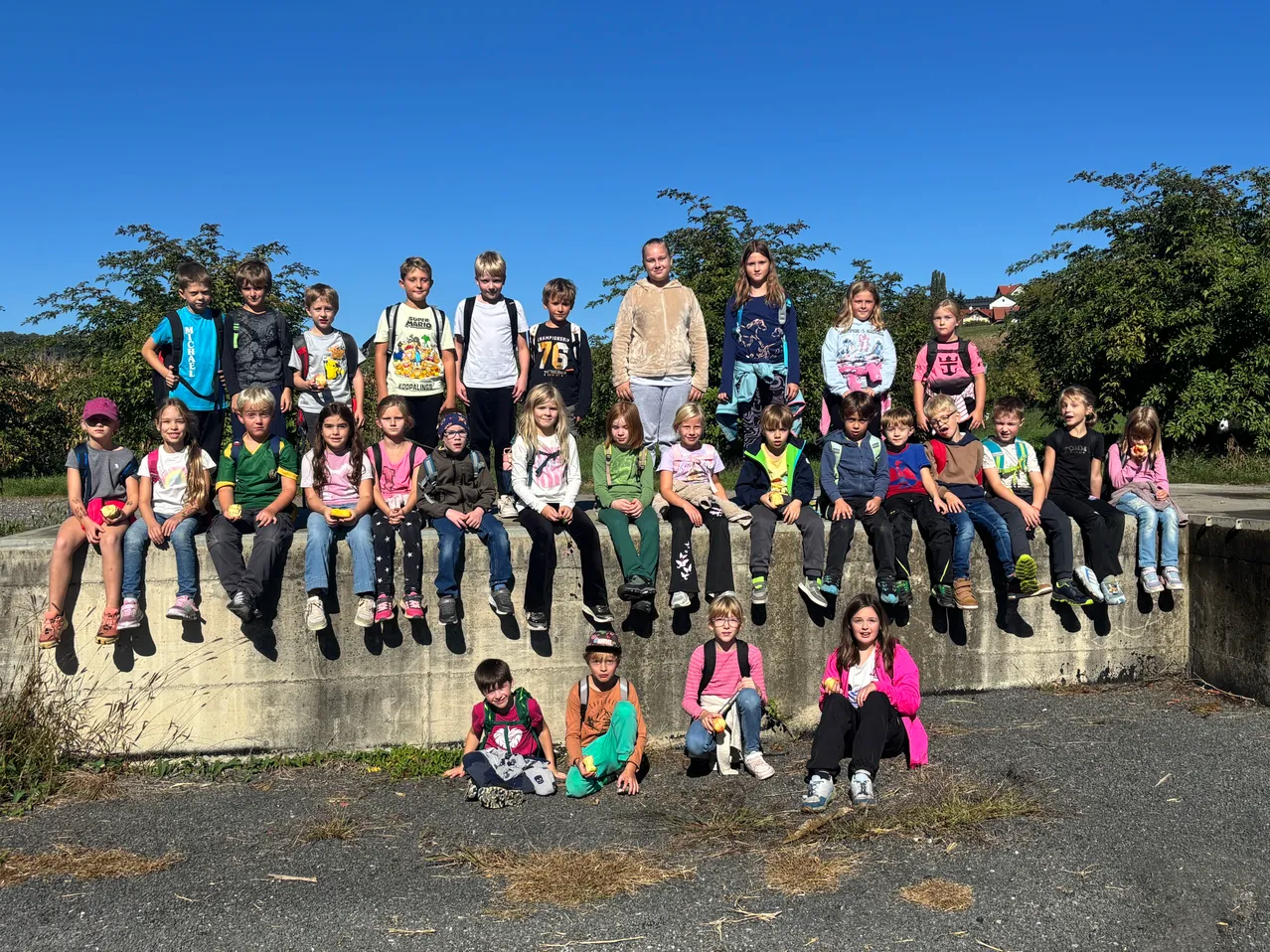 Eine Gruppe von Kindern, einige mit Rucksäcken, sitzen auf einer Betonbarriere und posieren für ein Foto. Die Kinder tragen verschiedene farbige Hemden und Turnschuhe. Der Hintergrund zeigt Bäume und einen klaren blauen Himmel.