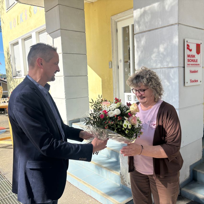 Ein Mann in einem Anzug überreicht einer Frau einen Blumenstrauß vor einem Gebäude mit einem Schild 'Musik Schule'.