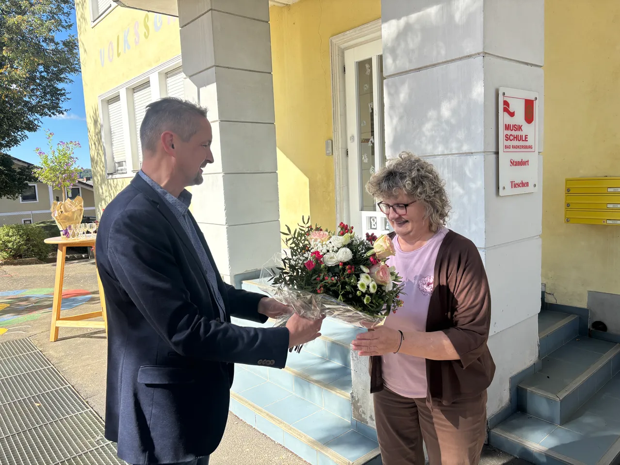 Ein Mann in einem Anzug überreicht einer Frau einen Blumenstrauß vor einem Gebäude mit einem Schild 'Musik Schule'.