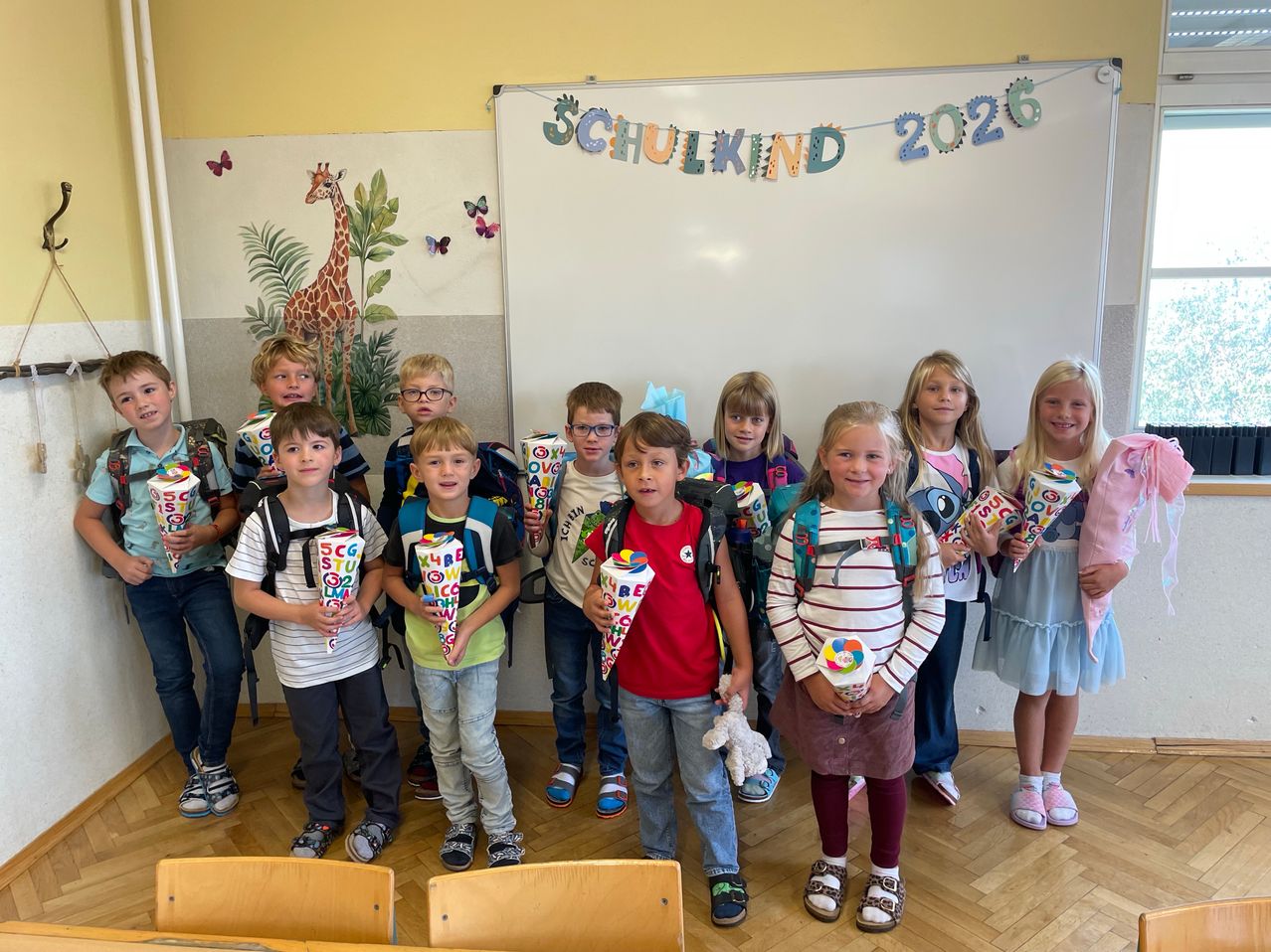 Eine Gruppe von Kindern im Klassenzimmer, alle mit Rucksäcken und Eistüten, vor einer Wand mit einem Banner, das Schulkind 2026 steht.