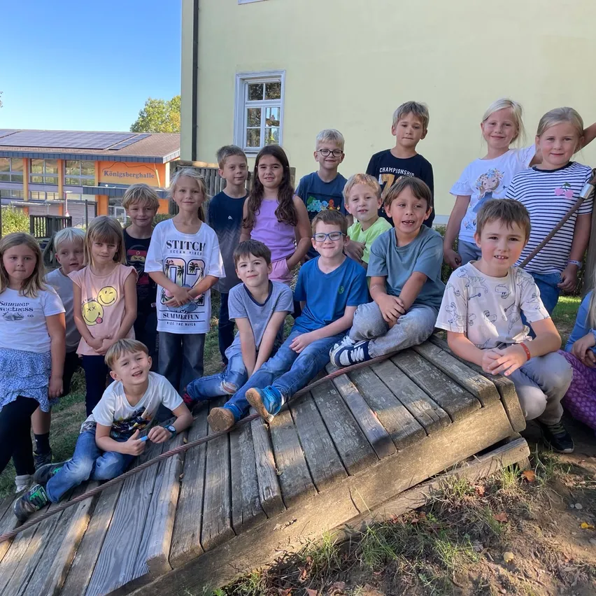 Eine Gruppe von Kindern in Freizeitkleidung posiert für ein Foto vor einem Gebäude. Einige sitzen auf einer Holzplanke, während andere hinter ihnen stehen.